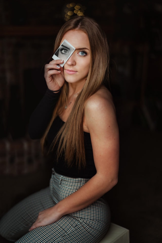 Creative senior picture with girl in dark room with black and white polaroid over eye