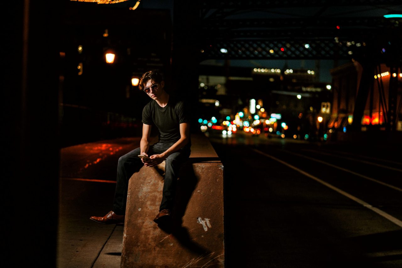 Senior guy at night sitting on the edge of a city street in downtown Minneapolis. Captured by Minnesota senior photographer Any Angle Photography.