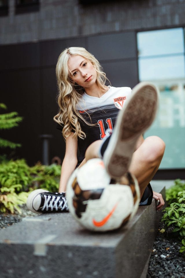 Minneapolis sports senior pictures in downtown Minneapolis in urban plaza with girl athlete in uniform sitting with foot resting on soccer ball. Captured by Minnesota senior photographer Any Angle Photography.