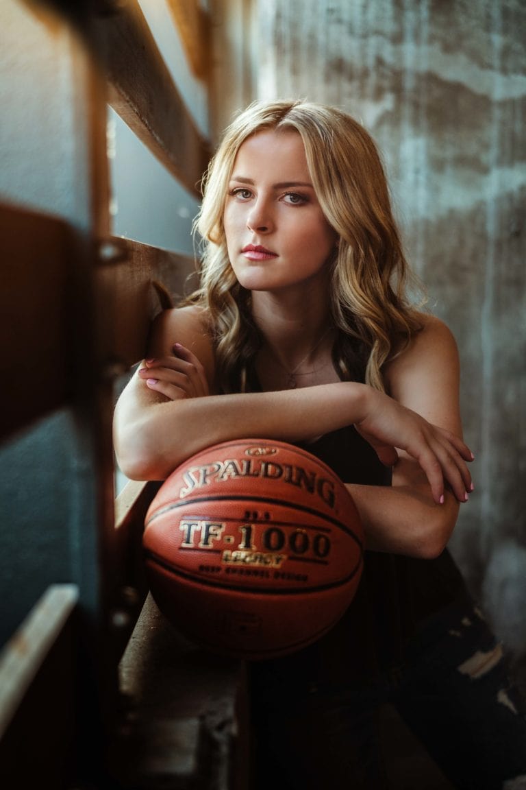 Minneapolis sports senior pictures with girl athlete holding basketball in parking garage leaning on ledge. Captured by Minnesota sports senior photographer Any Angle Photography.