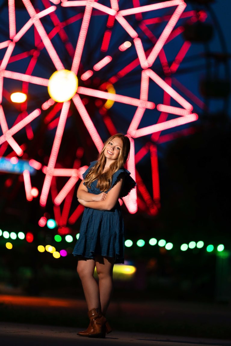 senior girl standing in front of a ferris wheel at night with bright lights