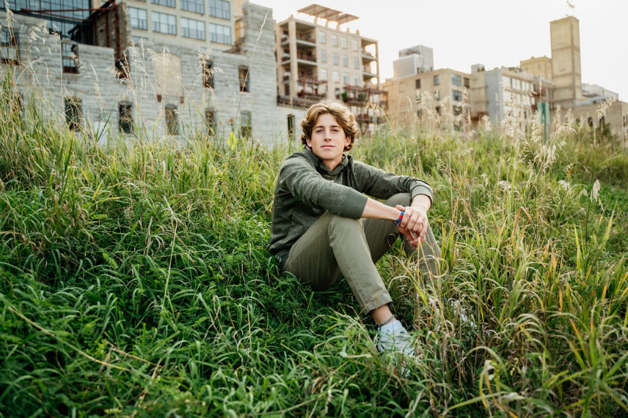 Senior pictures with guy sitting in tall grass in front of Mill City Museum in Minneapolis. Captured by Minneapolis senior photographer Any Angle Photography.