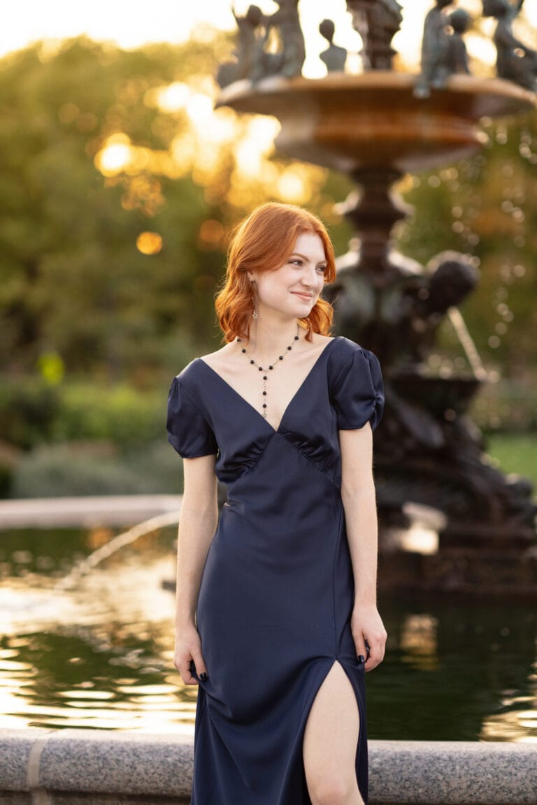 Nature senior pictures with girl with red hair in blue dress standing in front of a fountain in lyndale park rose garden in Minneapolis. Captured by Minneapolis senior photographer, Any Angle Photography.