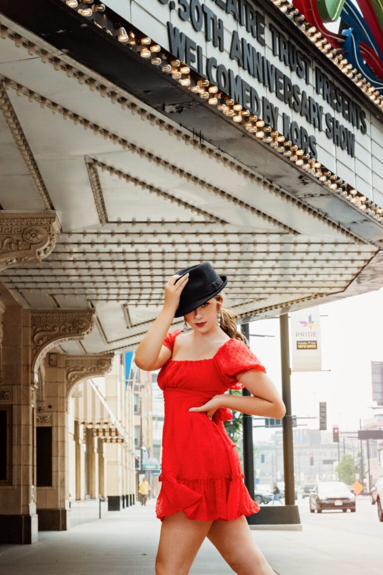 Senior pictures with girl in red dress and black fosse hat in front of theater in Minneapolis. Captured by Minnesota senior photographer, Any Angle Photography.