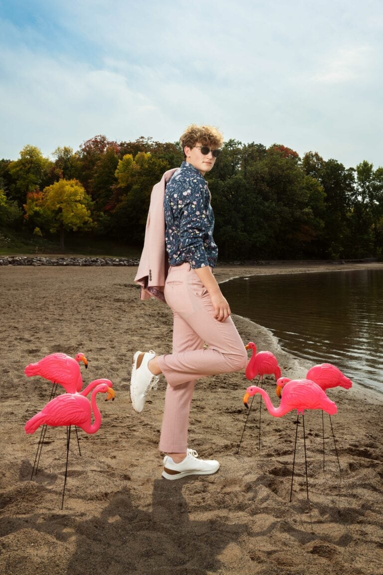 Guy standing on beach in pink suit surrounded by pink plastic flamingos. Captured by Minneapolis Senior Photographer, Any Angle Photography.